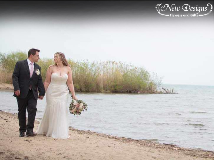 A bride and groom walk along the beach with their boquet and boutonniere in shades of white and pink